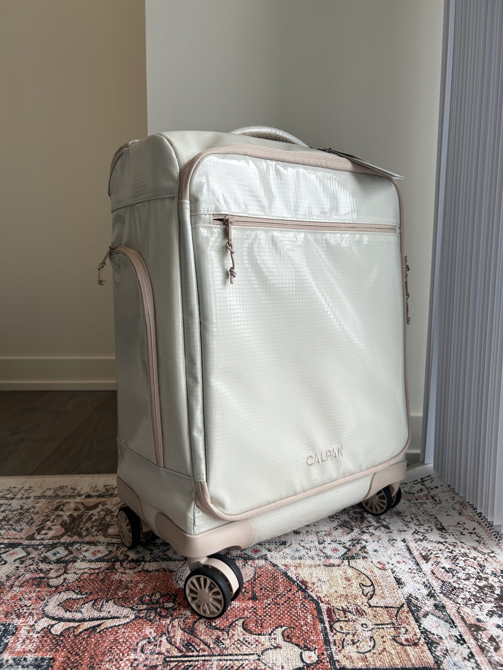 A light beige CALPAK rolling suitcase with four wheels stands upright on a patterned rug near a light-colored wall and a vertical panel.