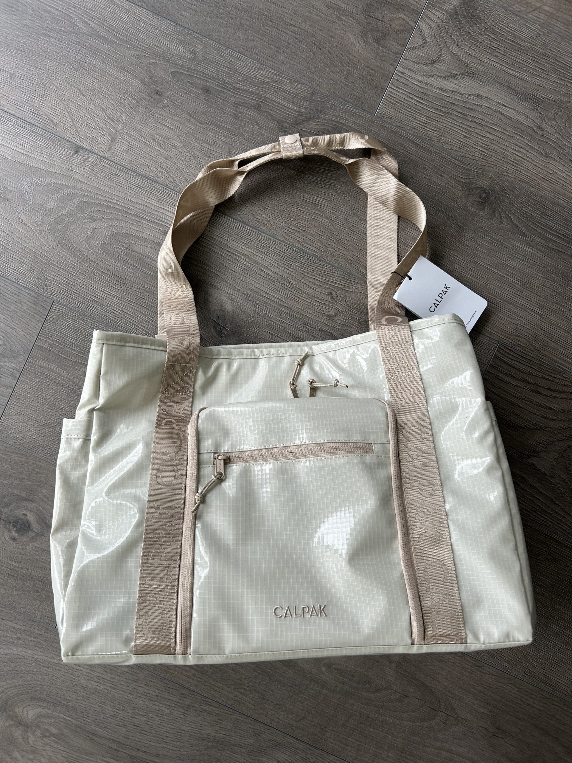 A cream-colored CALPAK tote bag with beige handles and a front zipper pocket is placed on a wooden floor.