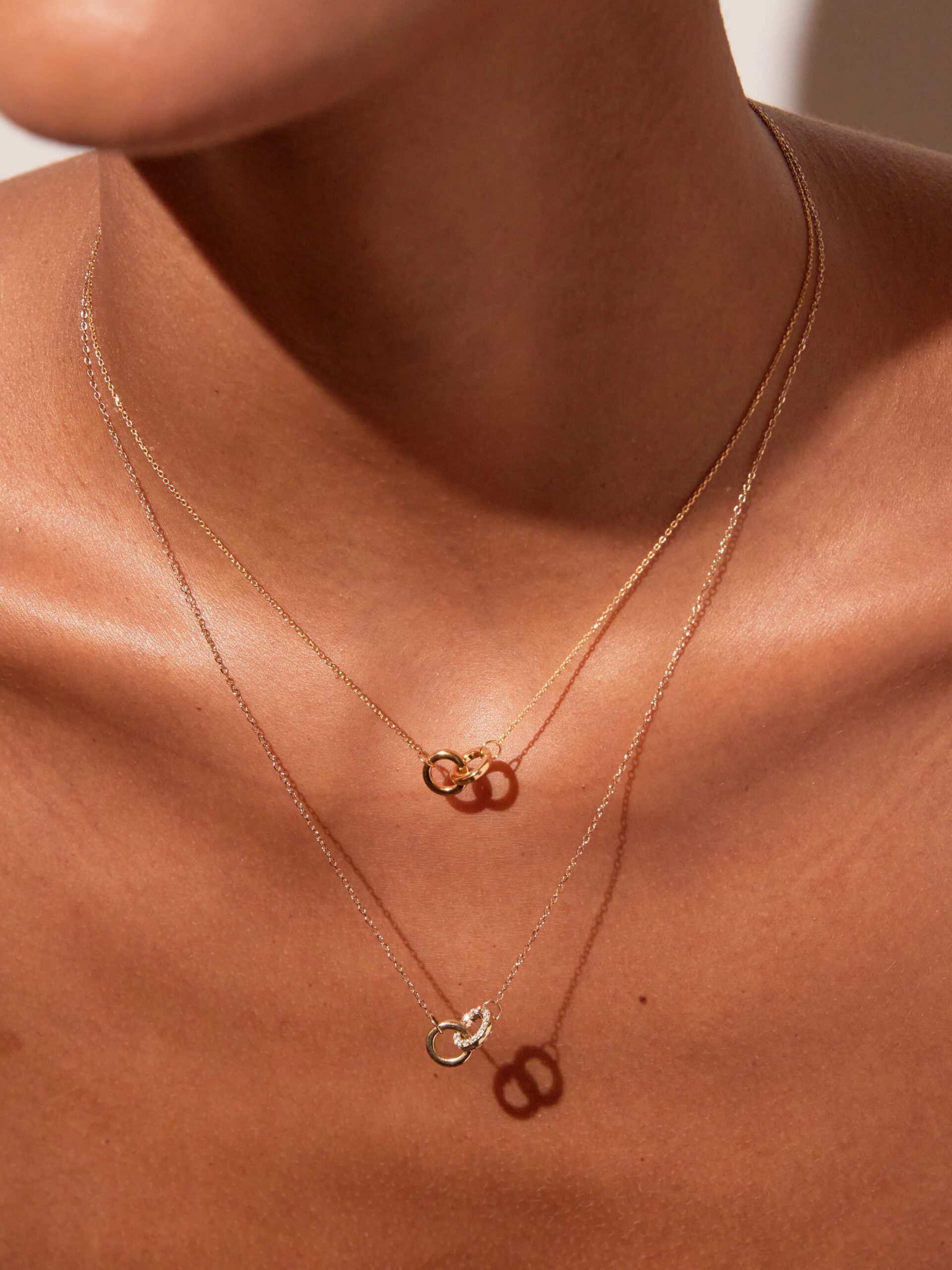 A close-up of a person's neck wearing two delicate gold necklaces, each with a small circular pendant, against a neutral background.