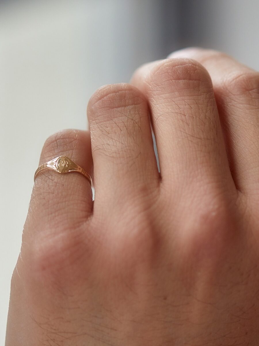 A hand with light skin wearing a thin gold ring with a small diamond-shaped design on the ring finger against a blurred background.