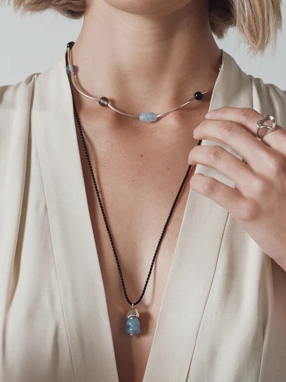 A person wearing a cream-colored v-neck top displays two necklaces with blue stone pendants and a silver ring on their finger.