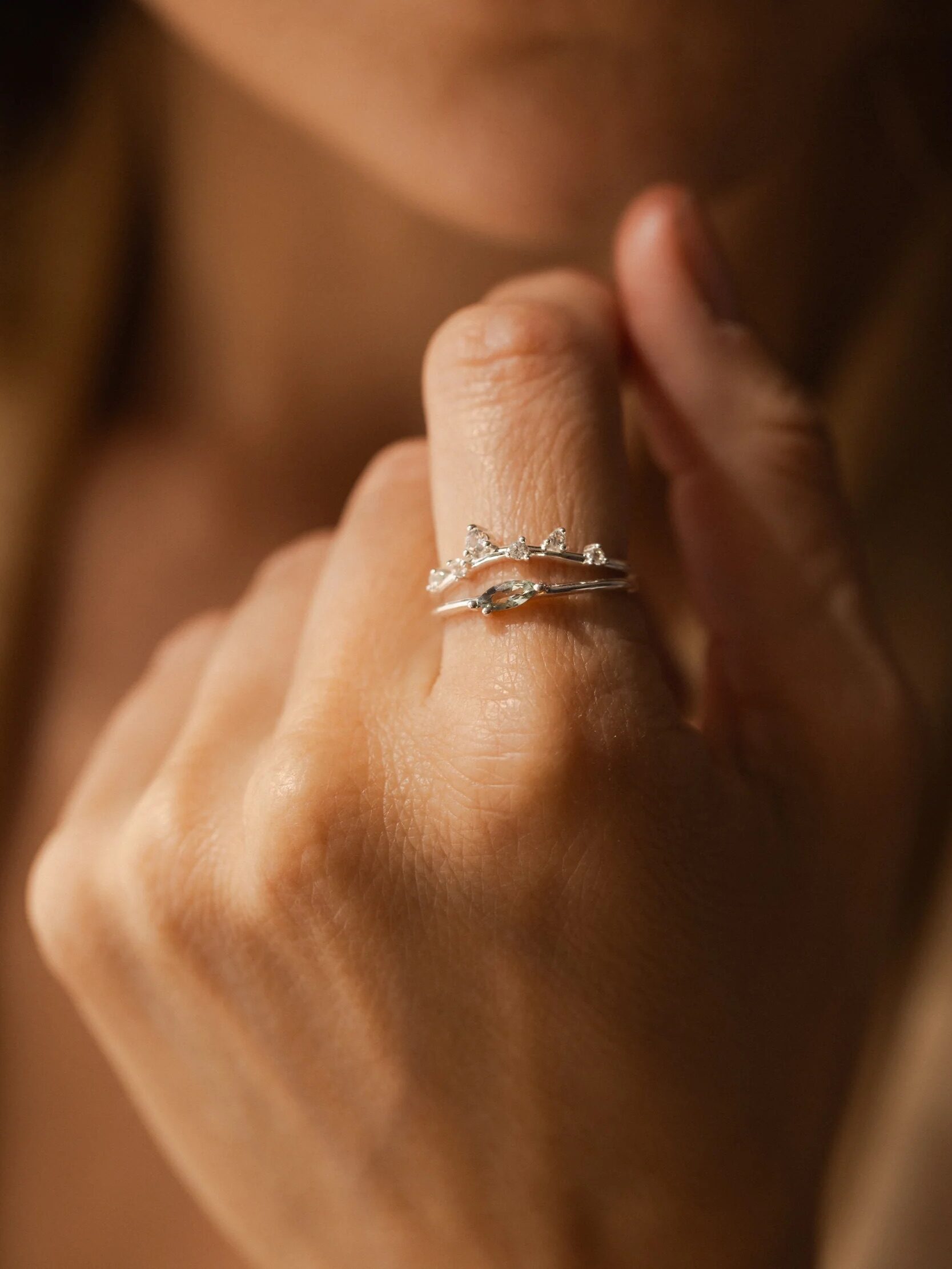 A hand with a light skin tone wears a silver ring featuring a wavy design and small gemstones, held close to the face.