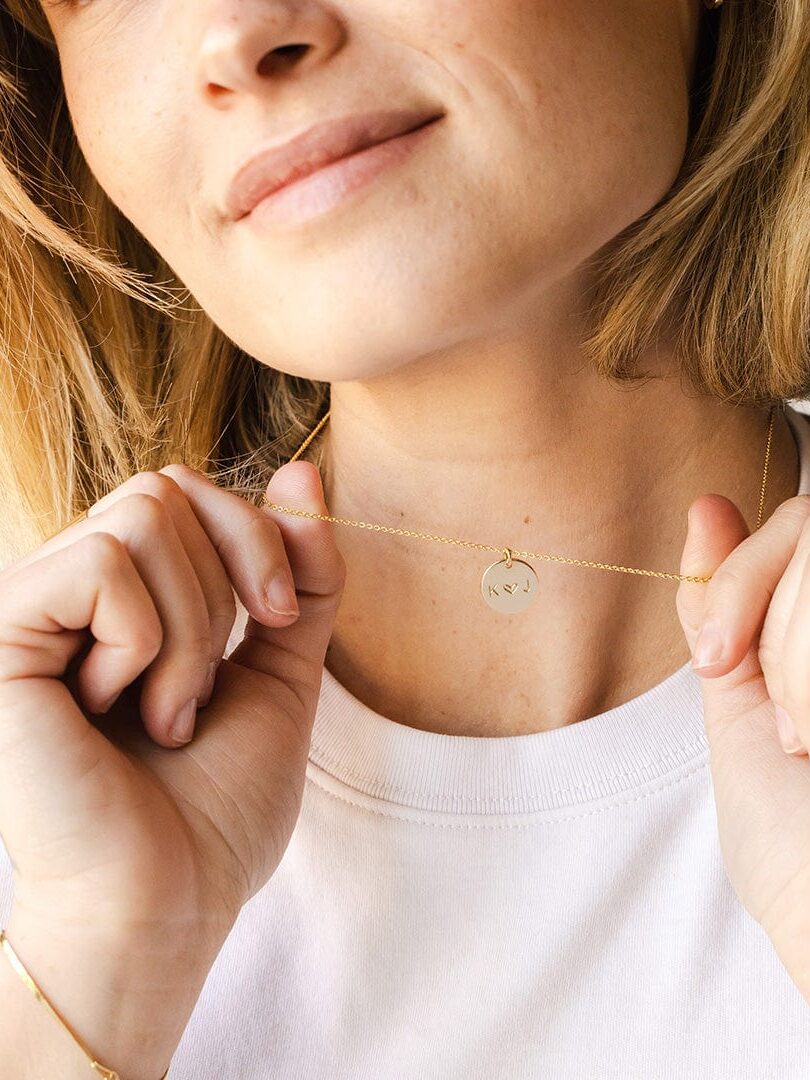 A woman wearing a white shirt holds a gold necklace with a round pendant engraved with the initials "RSV".