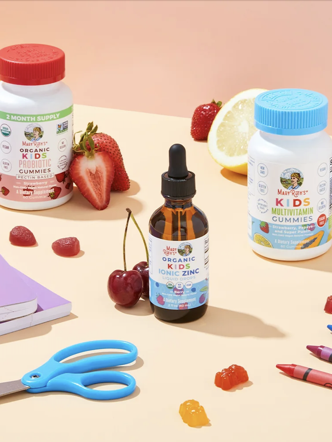 Three MaryRuth’s kids supplement products, fruits, scattered gummies, crayons, a notebook, scissors, and a pencil case are arranged on a pastel surface.