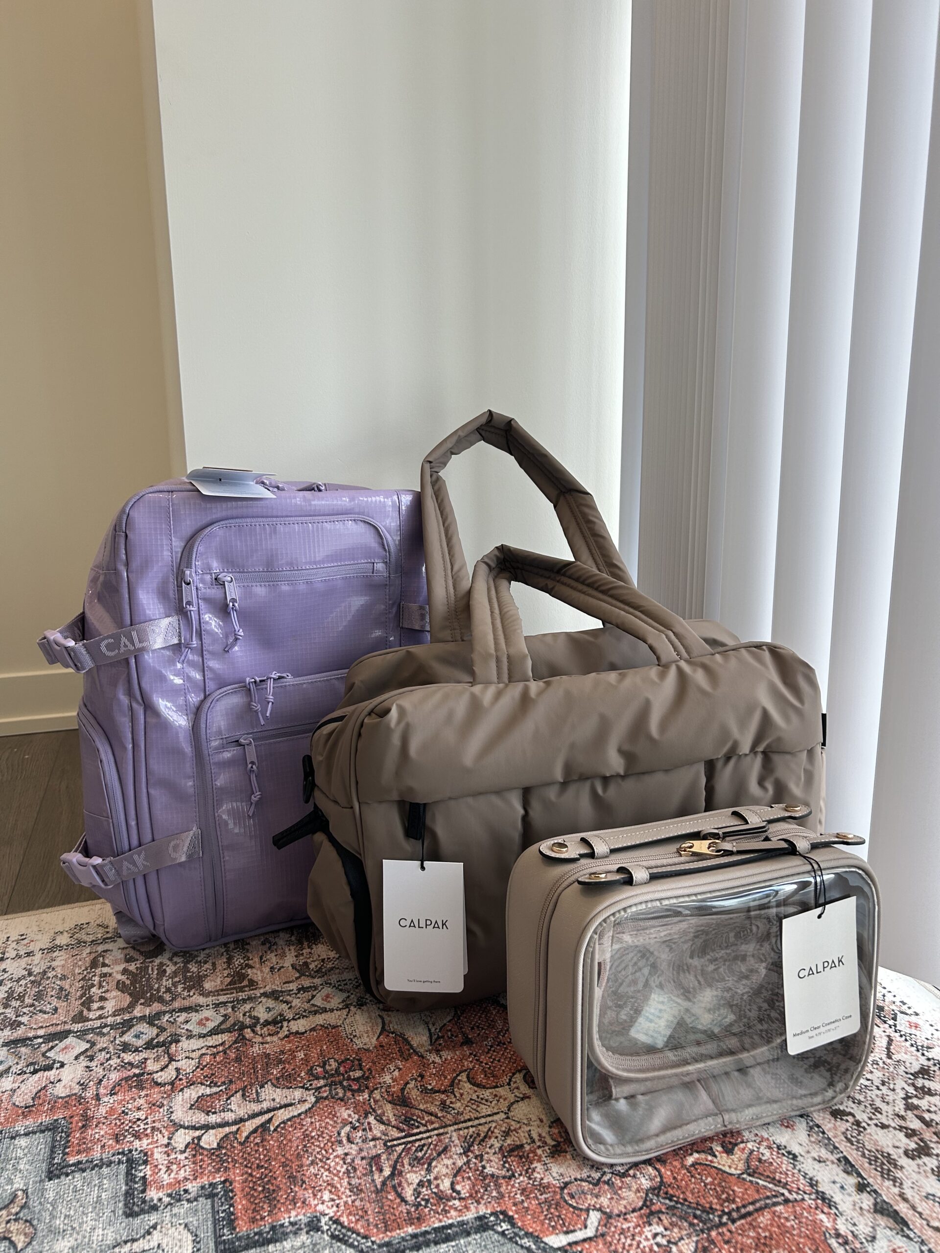 Three CALPAK travel bags—a lavender backpack, a taupe duffel, and a clear toiletry case—are arranged on a patterned rug near a window with vertical blinds.