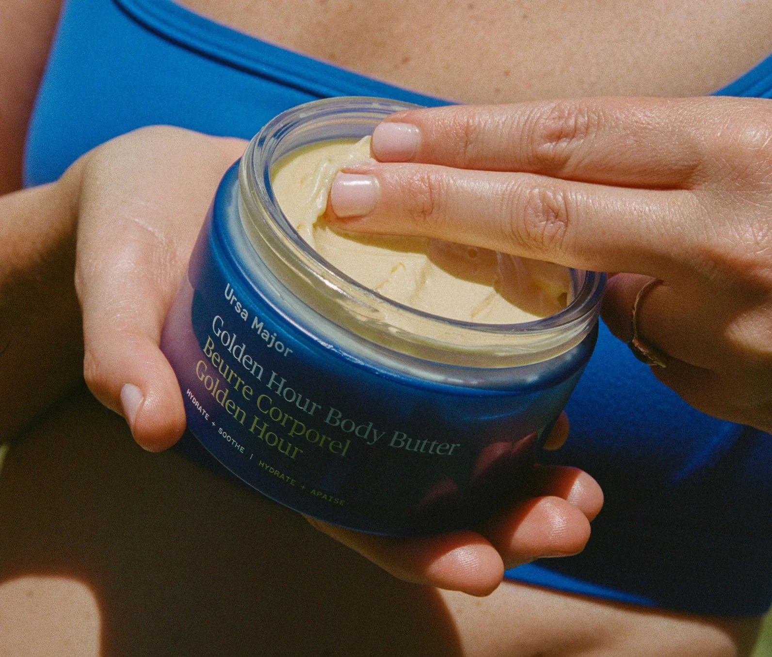 A person dips fingers into a jar labeled "Golden Hour Body Butter" by Ursa Major, the label visible and wearing a blue top—perfect for treating yourself during the Labor Day Sale.