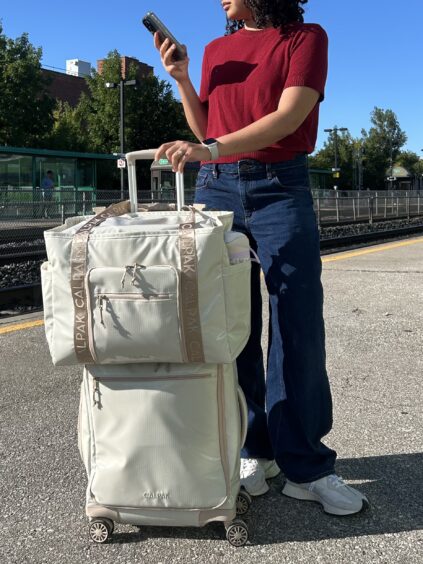 A person in a red shirt and blue jeans stands on a train platform, holding a phone and a large beige rolling suitcase with a matching bag stacked on top.