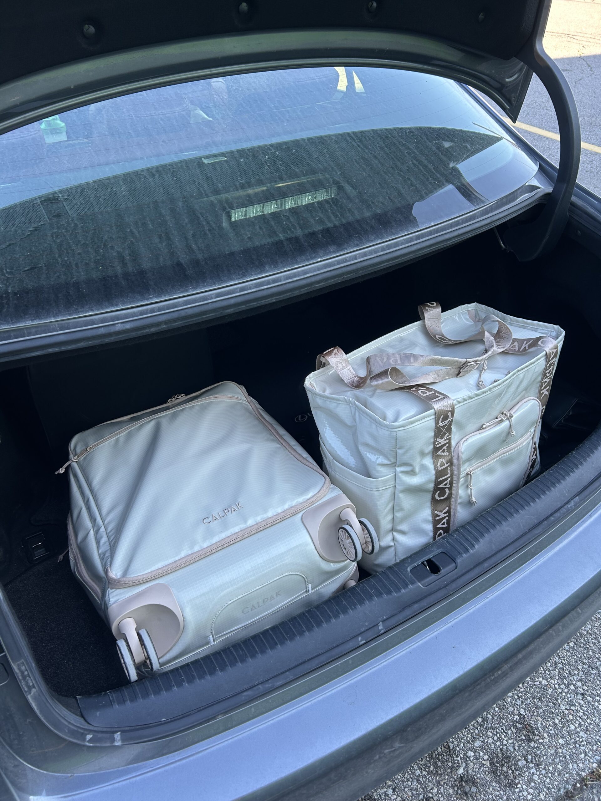 Two light-colored travel bags are placed in the trunk of a car, one with wheels and one with straps, both labeled "CLARA K.