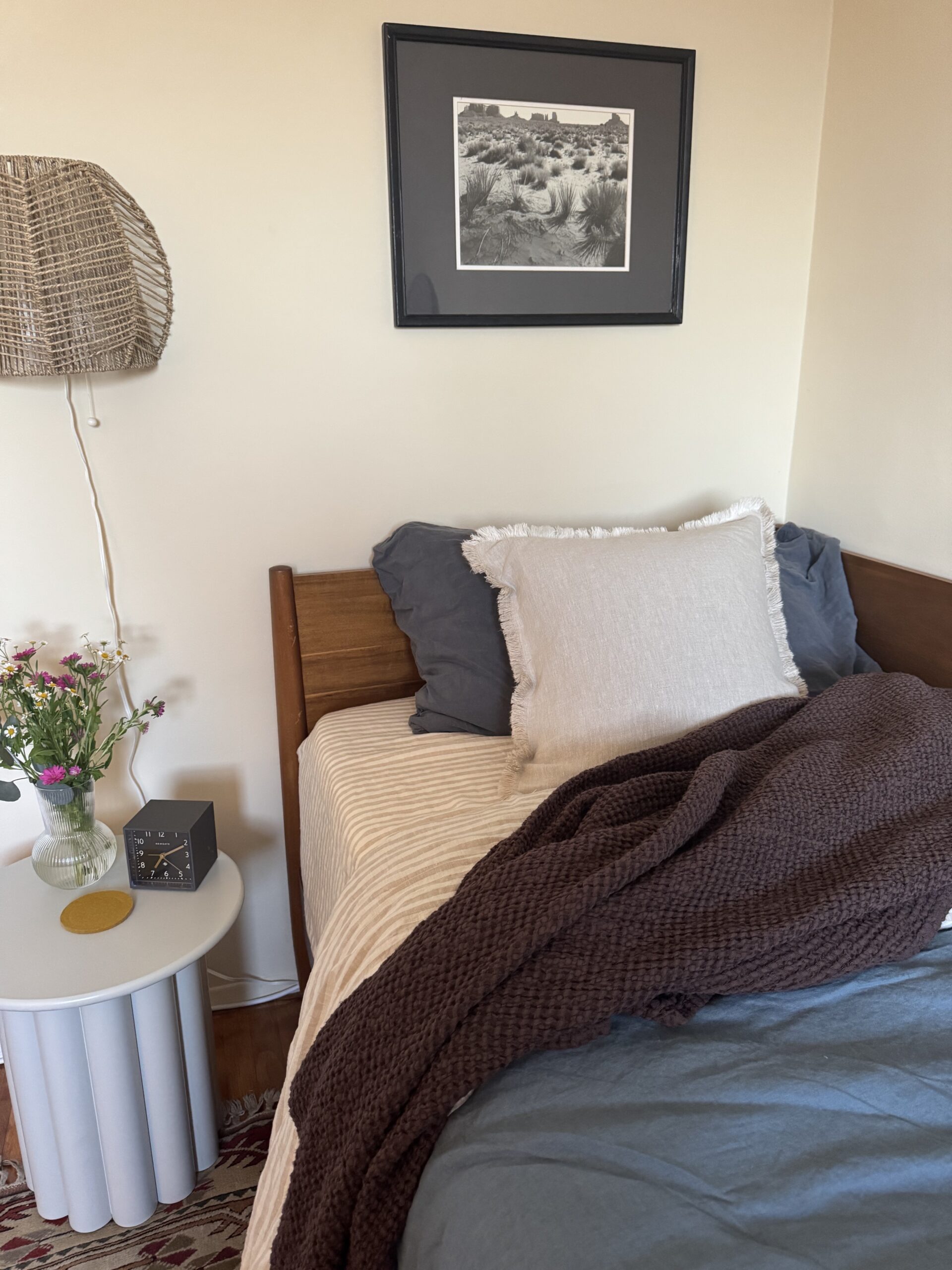 A neatly made bed with neutral linens sits beside a round white nightstand holding flowers, a clock, and a coaster; a framed black-and-white photo hangs above.