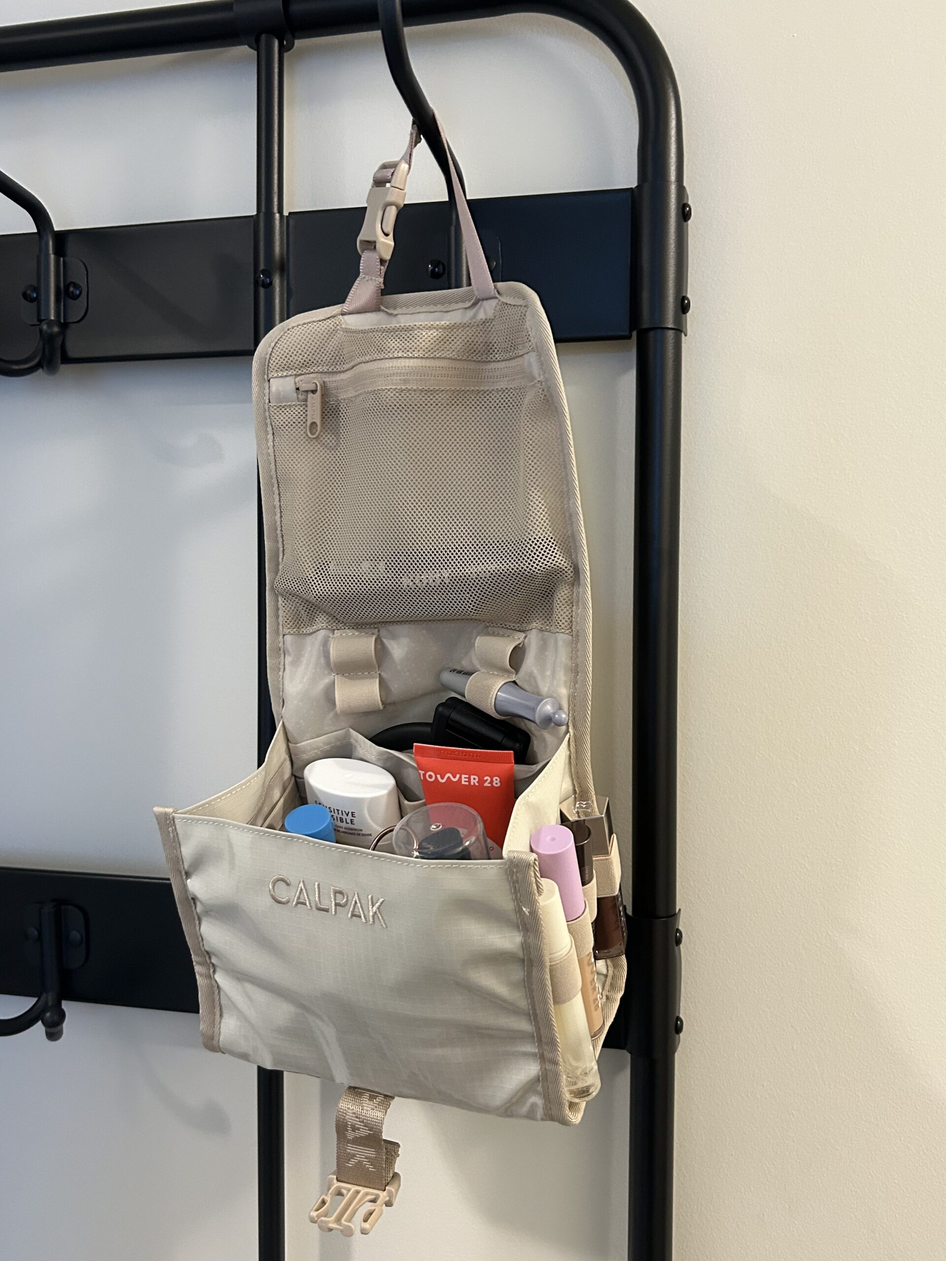 A beige hanging toiletry bag labeled "CALPAK" is open, displaying various skincare and makeup products organized in compartments.