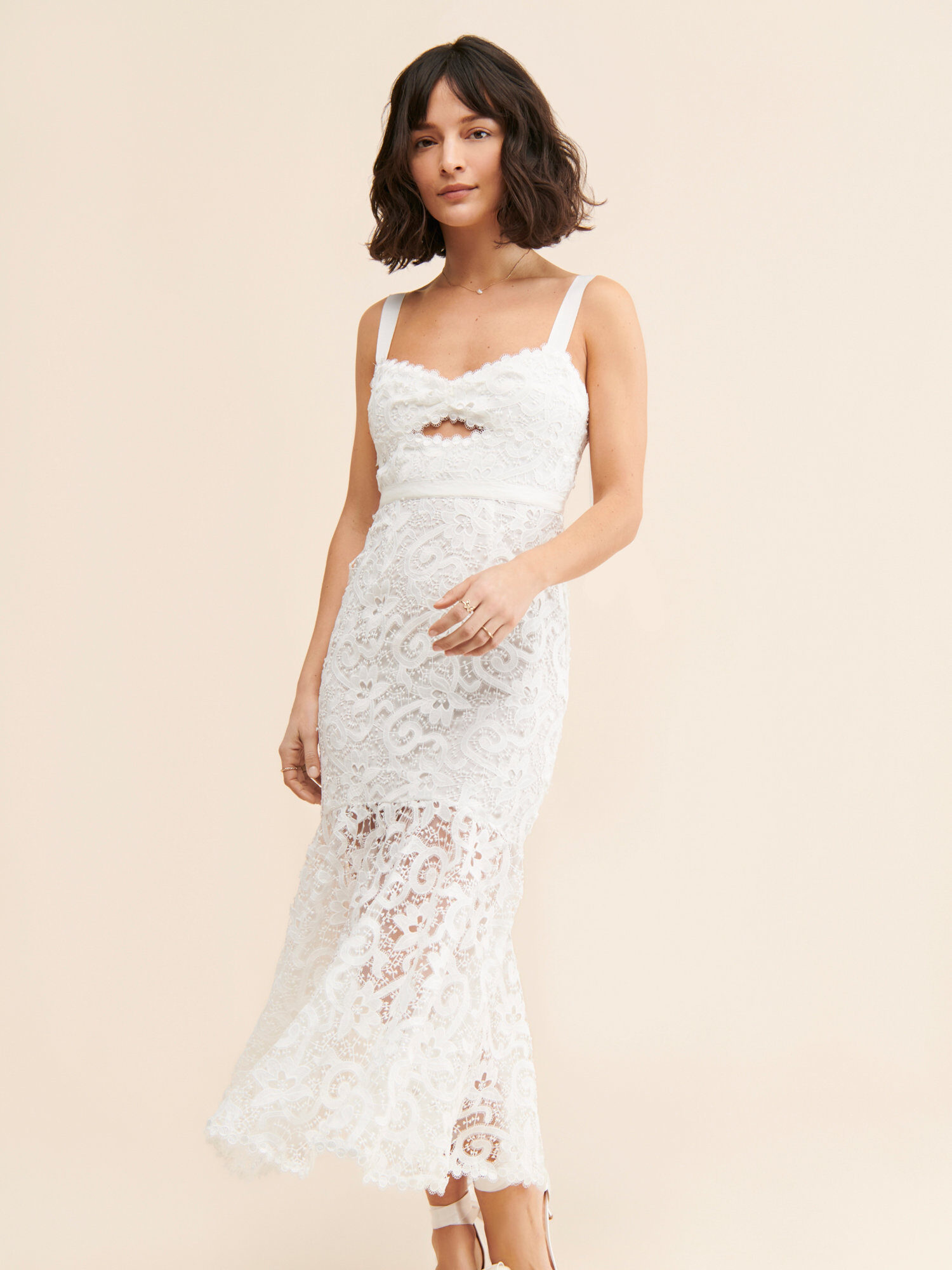 A woman wearing a white lace sleeveless dress with a cutout detail and bow-embellished heeled sandals, standing against a plain beige background.