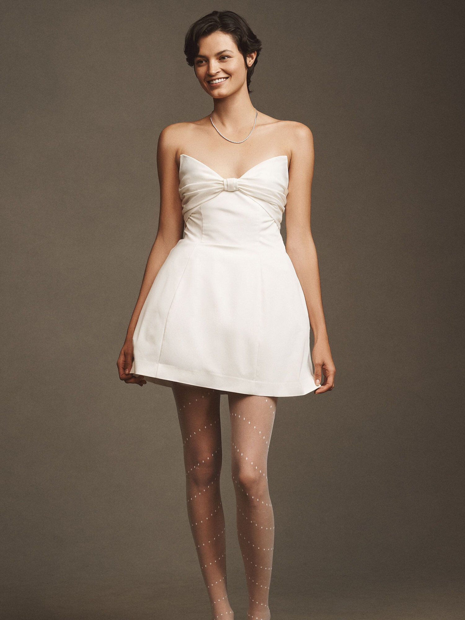 A woman stands facing forward, wearing a strapless white mini dress with a bow detail, sheer dotted tights, and pointed shoes against a plain background.