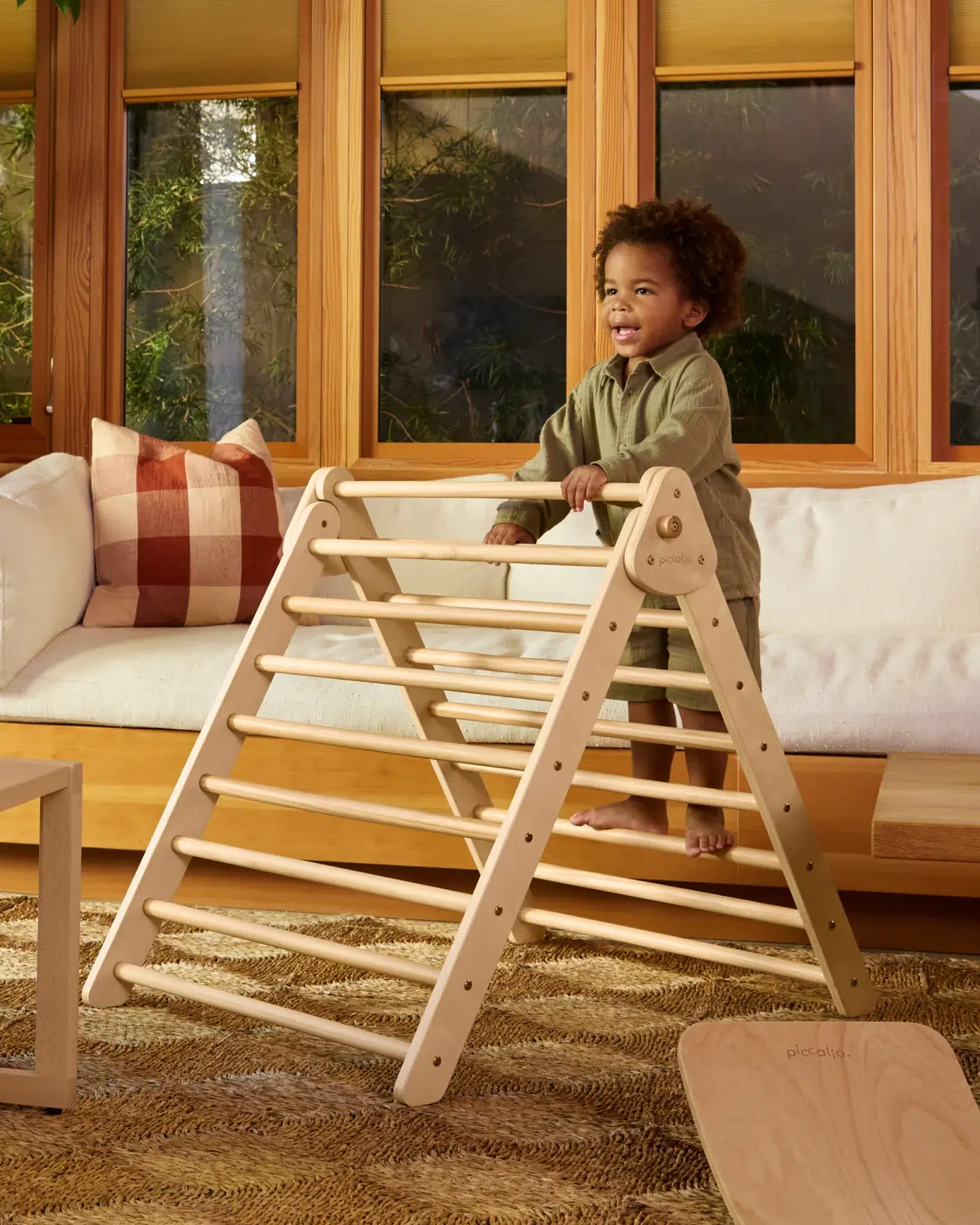 A young child stands and plays on a wooden climbing triangle in a sunlit living room with wooden accents and a white sofa.