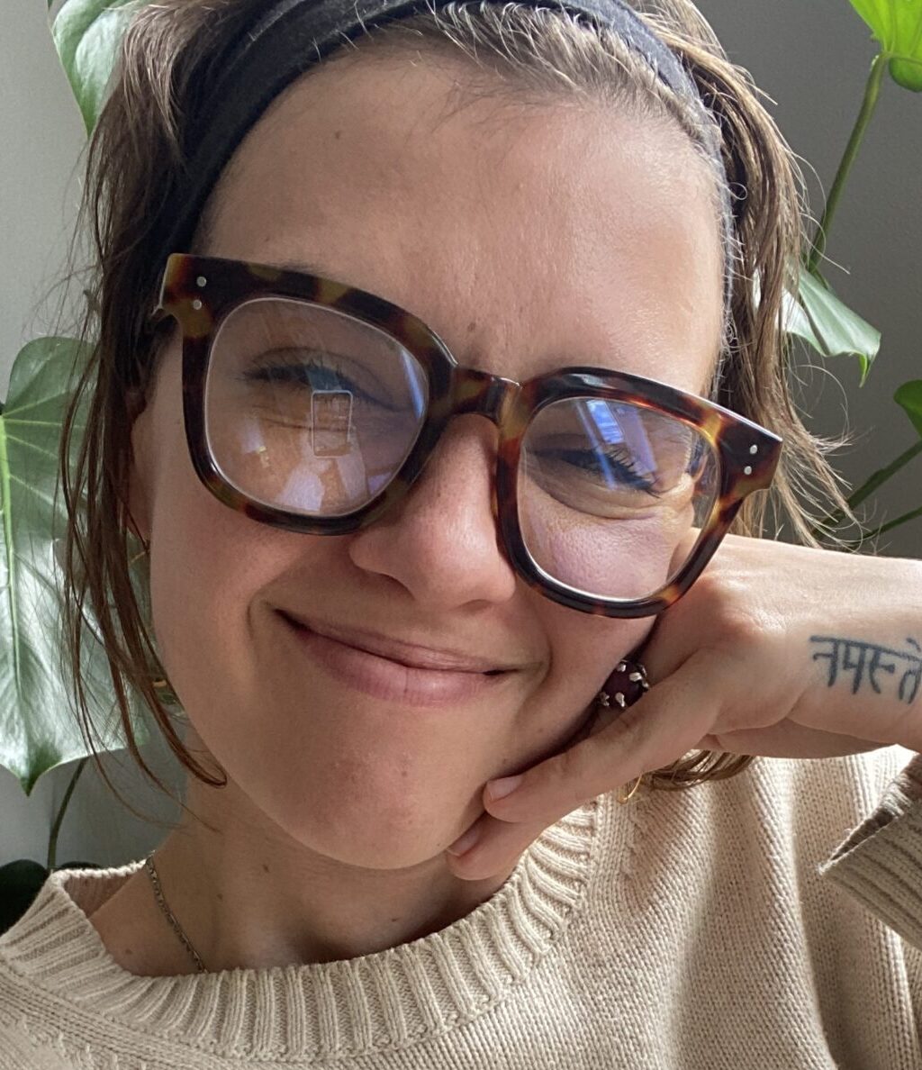 Person wearing large tortoiseshell glasses and a beige sweater smiles with their chin resting on their hand; a "Namaste" tattoo is visible on their wrist.