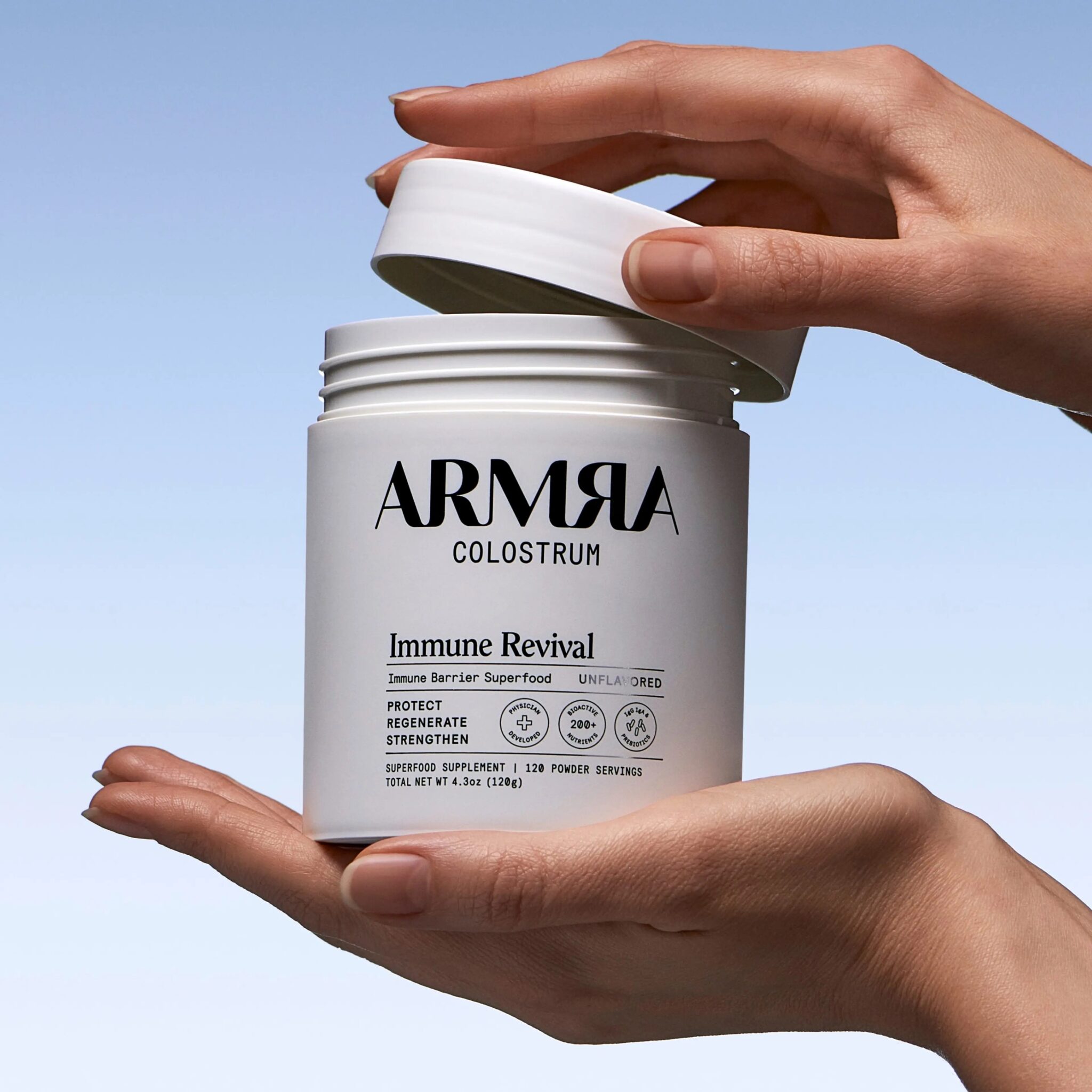 A person opens a white container labeled "ARMRA Colostrum Immune Revival" against a light blue background.