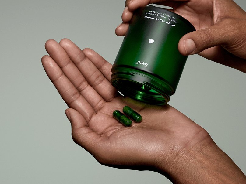 A person pours two green capsules from a dark green bottle into their palm against a plain background.