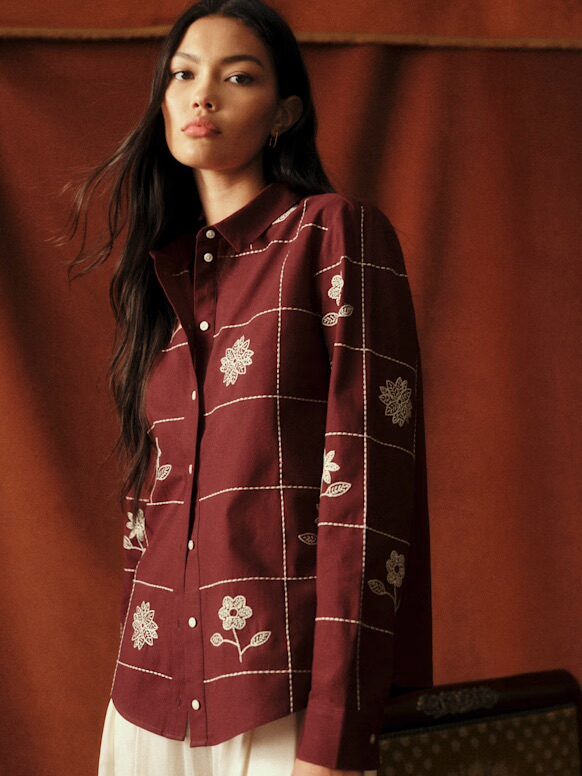 Woman wearing a maroon button-up shirt with white floral embroidery and cream-colored pants, standing against a brown background.