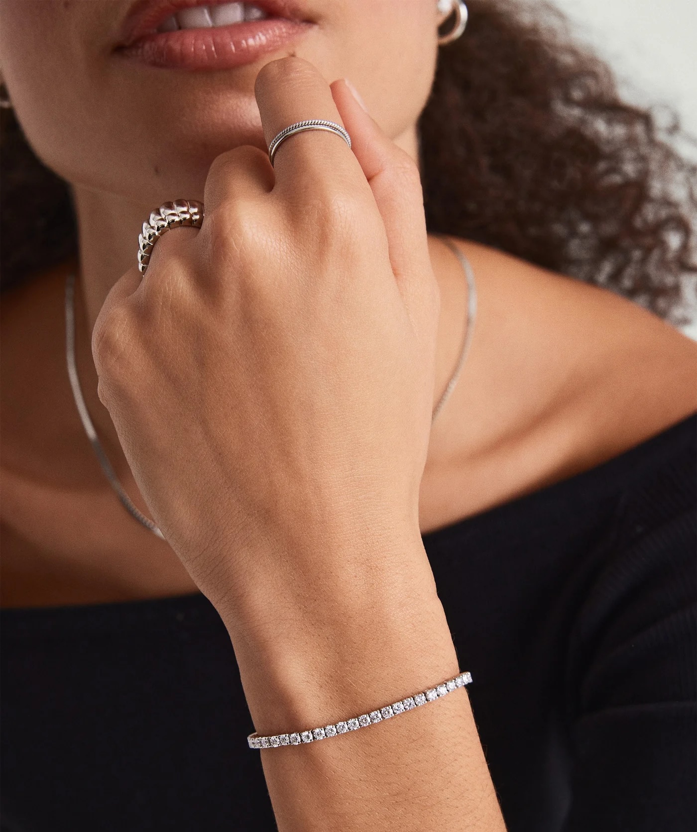 A person wearing a black top displays a silver necklace, two rings, and a tennis bracelet with clear stones on their hand and wrist.