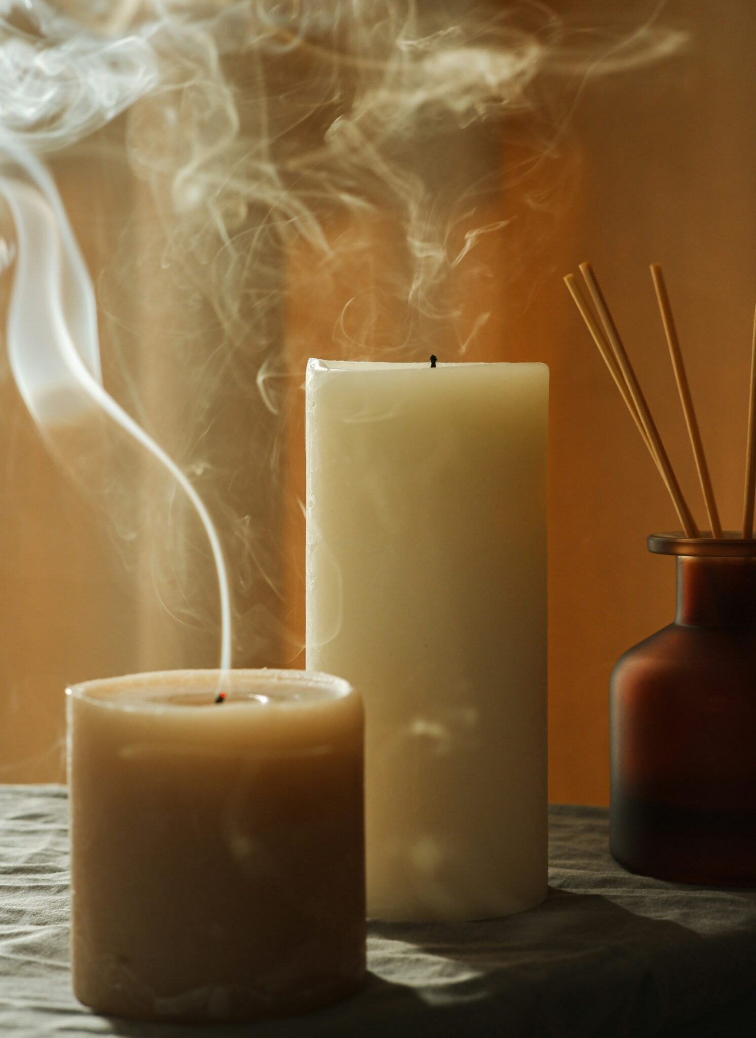 Two unlit pillar candles, one with visible smoke rising from its wick, stand on a fabric surface next to a brown glass diffuser bottle with reed sticks.
