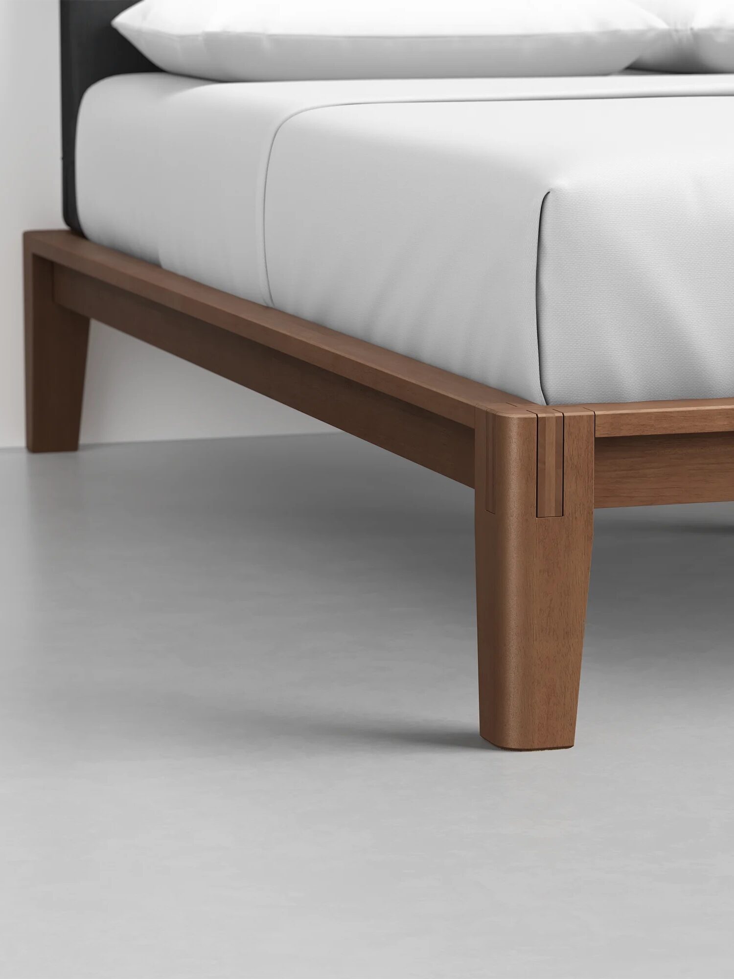 Close-up view of a wooden bed frame with tapered legs and a white mattress, placed on a smooth, light gray floor.