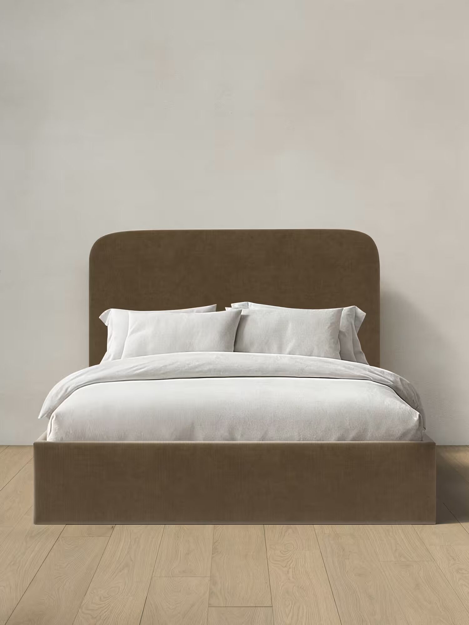 A brown upholstered bed frame with a tall, rounded headboard is made up with white bedding and pillows, positioned against a plain light-colored wall on a wood floor.