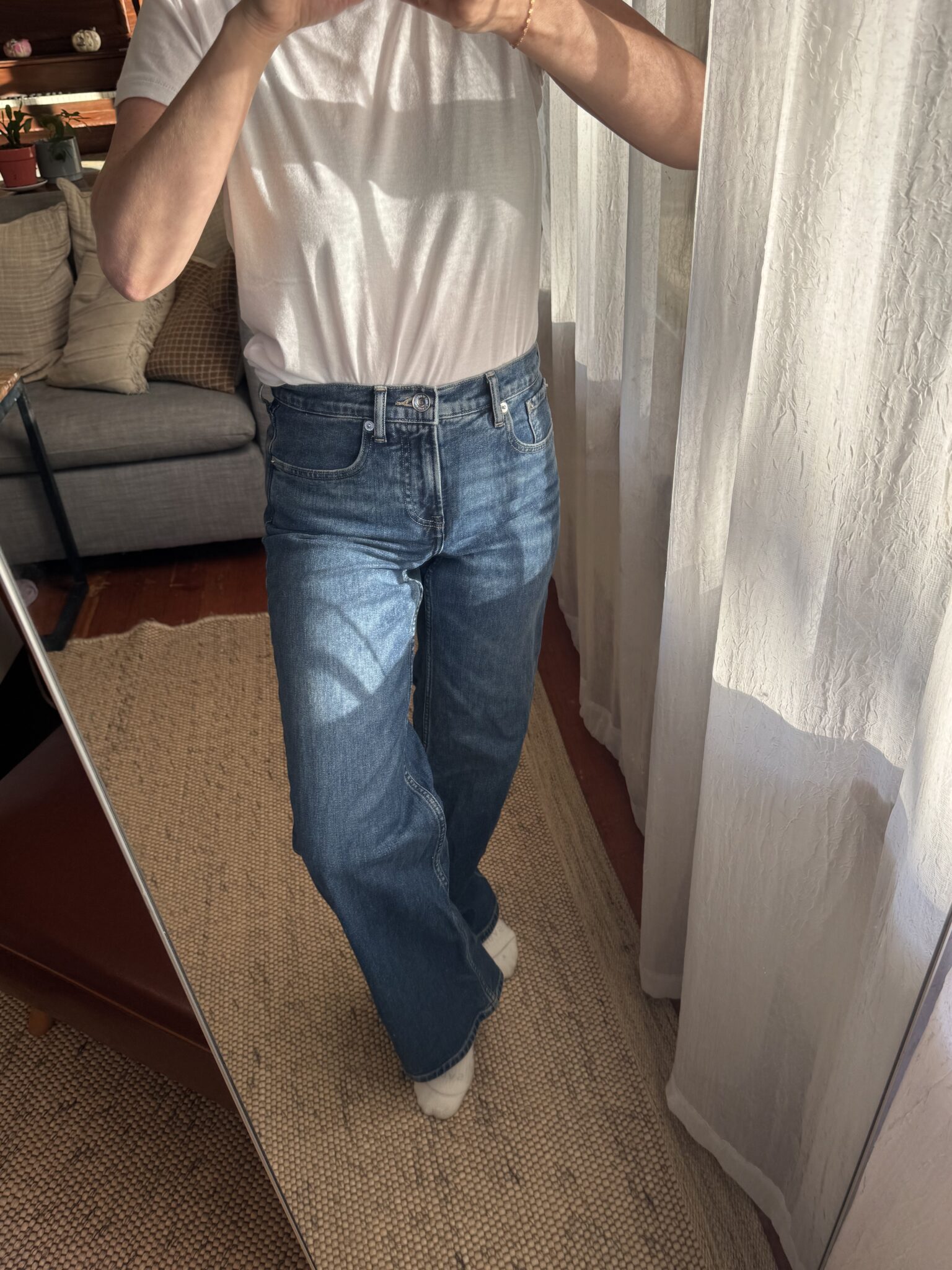 Person wearing a white t-shirt, blue high-waisted jeans, and white socks stands on a neutral rug, taking a mirror selfie in a well-lit room.