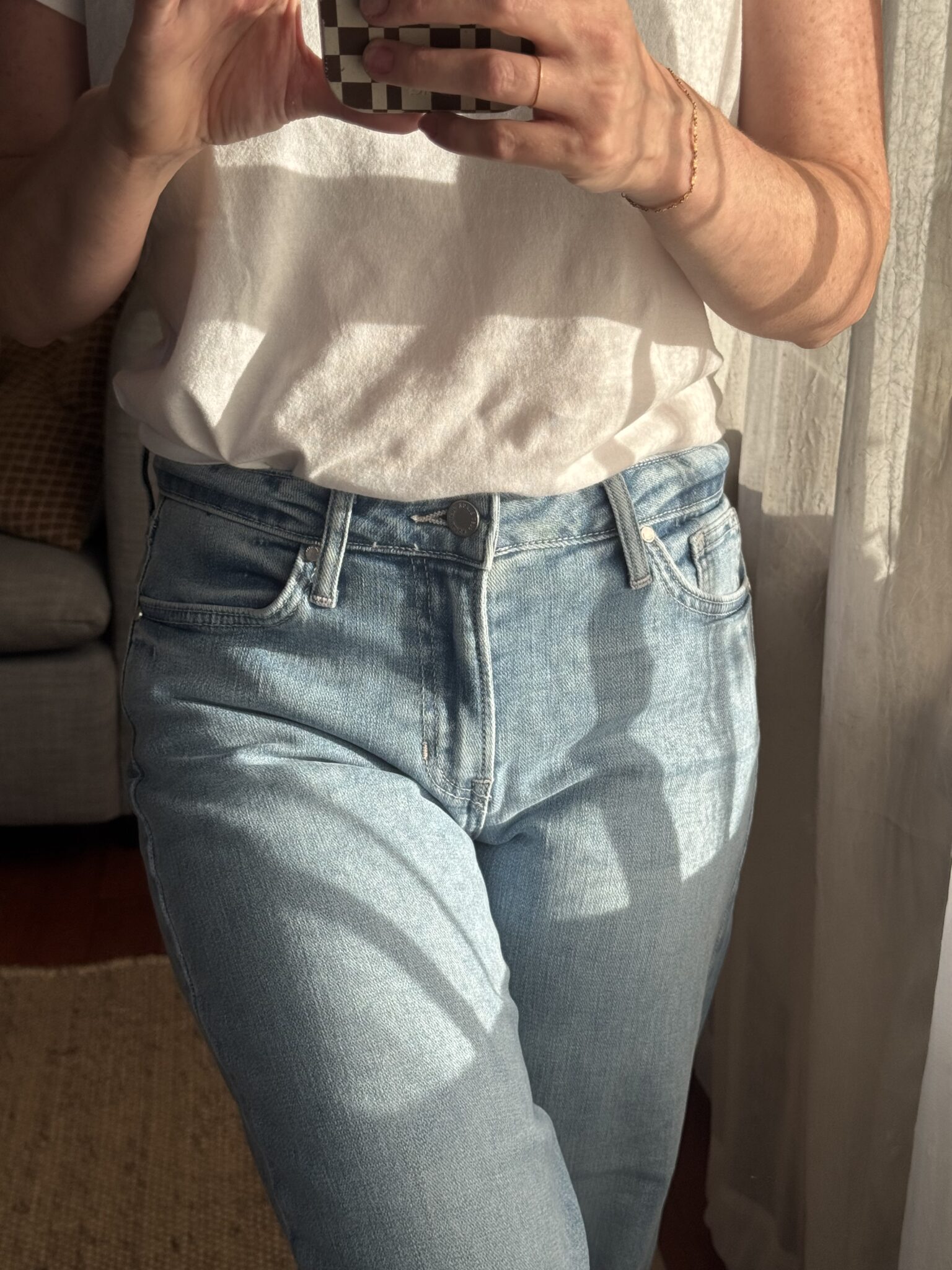 Person wearing a white sleeveless top tucked into light blue jeans, holding a phone to take a photo in natural sunlight.