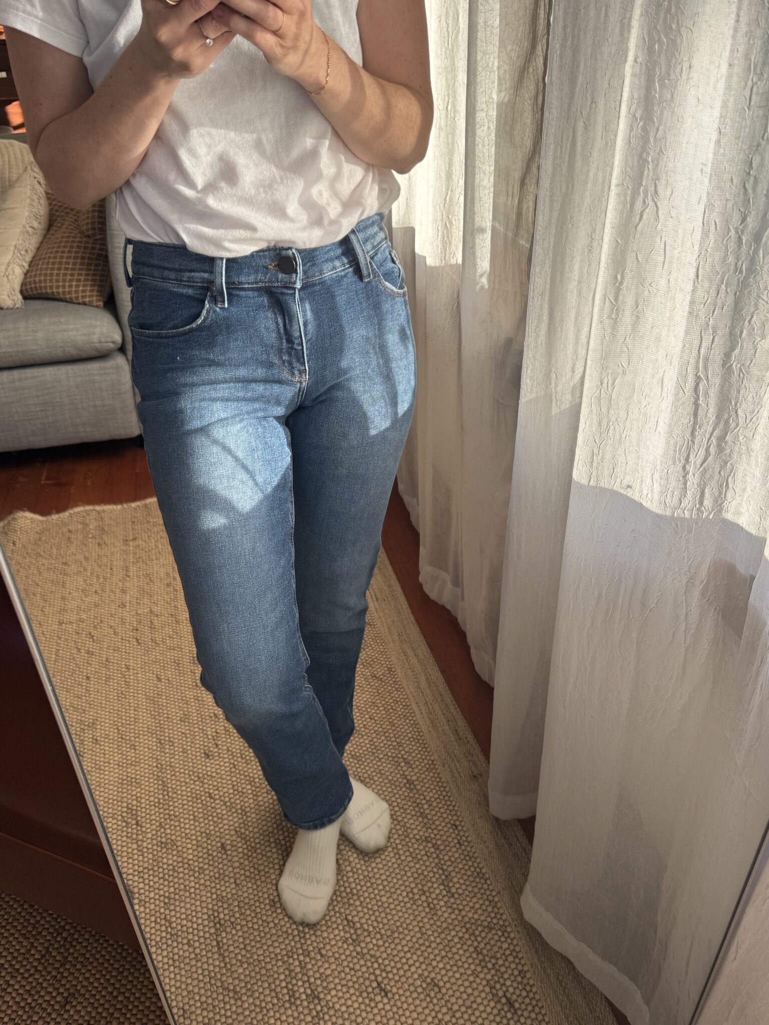 Person wearing a white t-shirt, blue jeans, and white socks stands in front of a mirror, taking a photo. Light-colored curtains and a woven rug are visible in the background.
