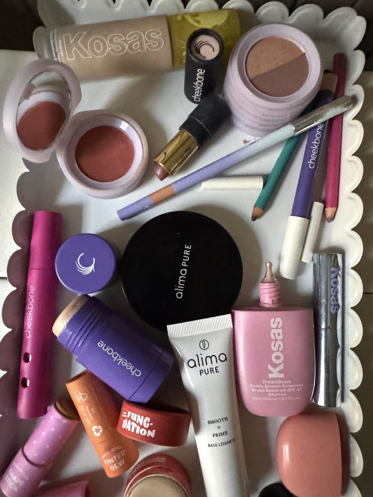 A variety of makeup products, including lipsticks, blushes, pencils, and creams, are arranged on a white scalloped tray. Brands such as Kosas, Cheekbone, and Alima Pure are visible.