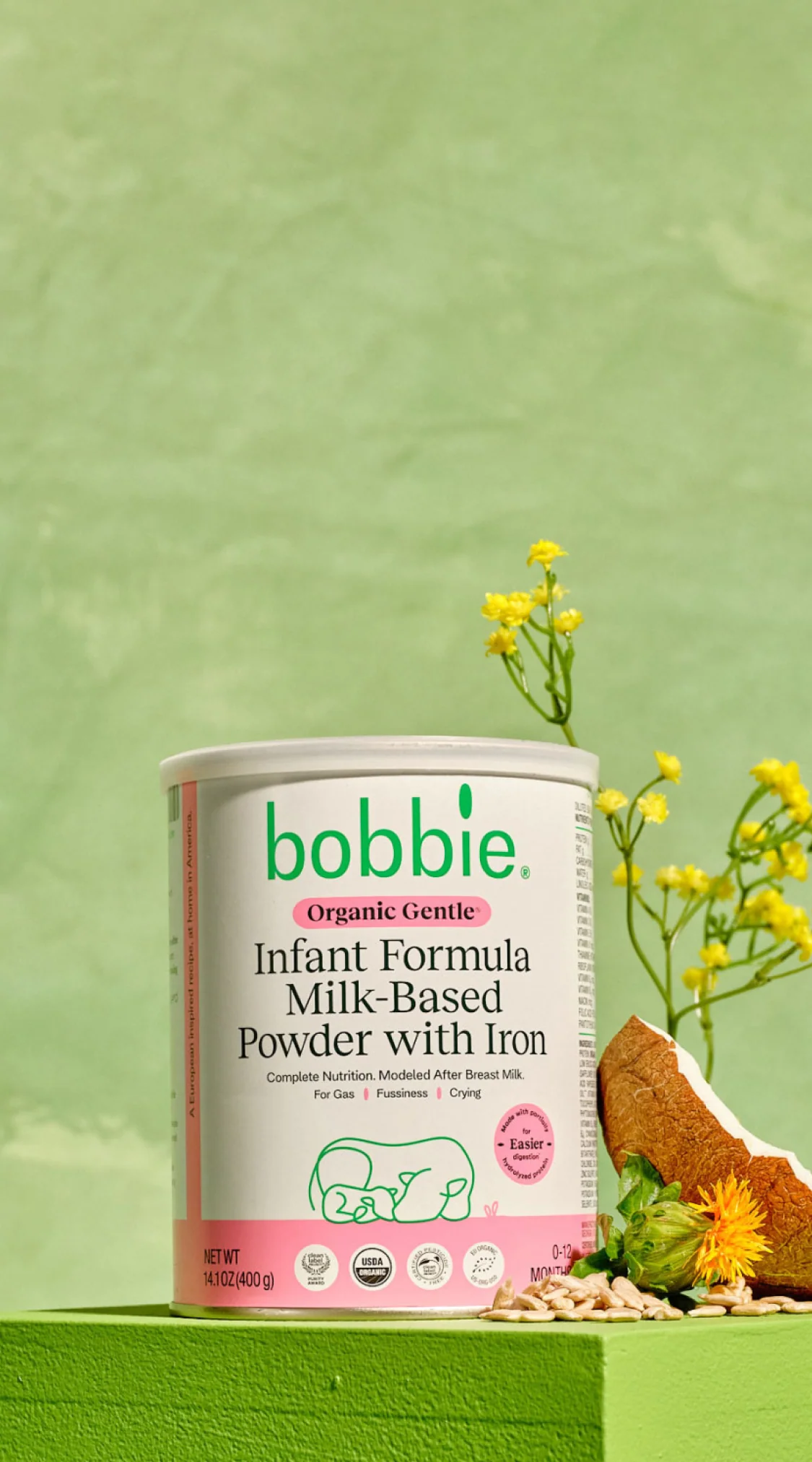 A canister of Bobbie Organic Gentle Infant Formula Milk-Based Powder with Iron stands on a green surface, next to yellow flowers and grains, against a green background.