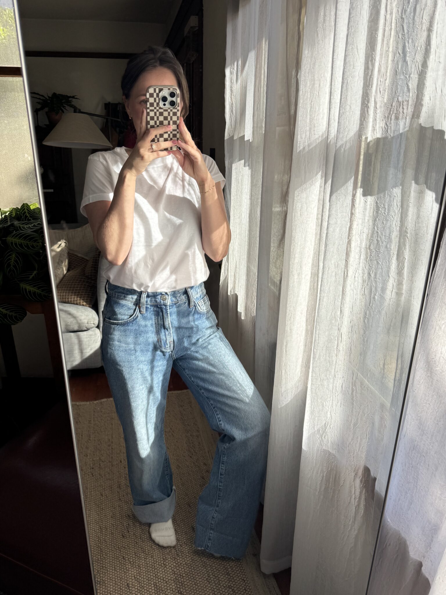 Person in a white t-shirt, blue wide-leg jeans, and white socks stands indoors, taking a mirror selfie with a phone in a checkered case. Sunlight filters through sheer curtains.