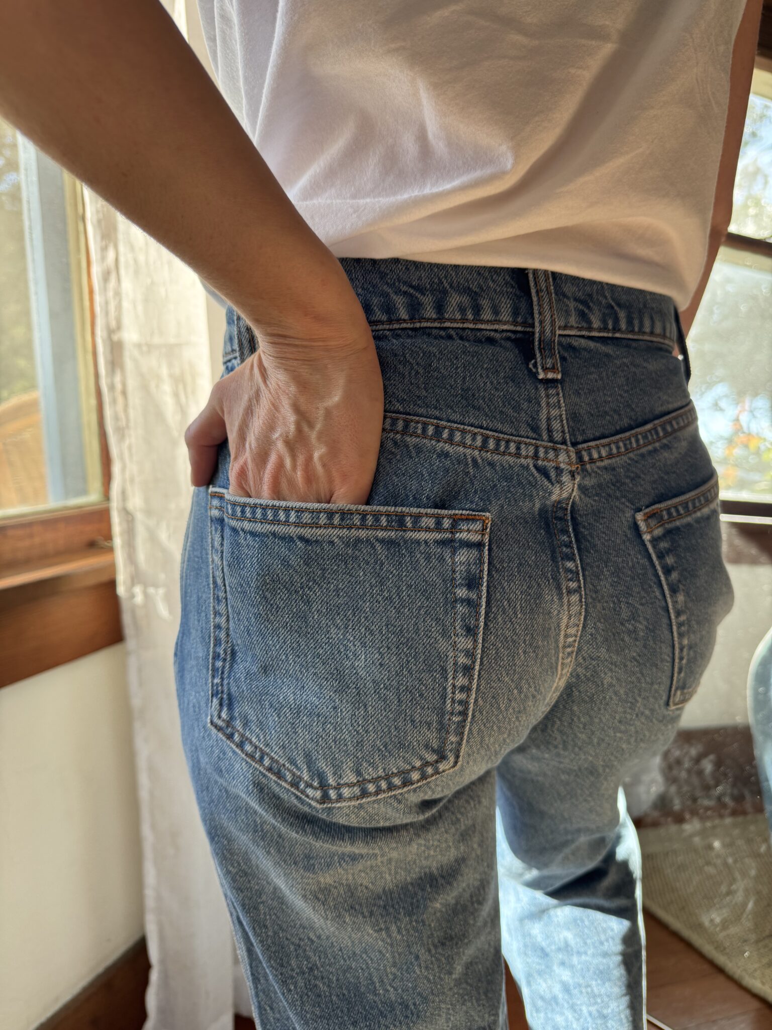 Person in blue jeans and a white shirt stands indoors with their right hand in the back pocket, near a window with sunlight coming in.