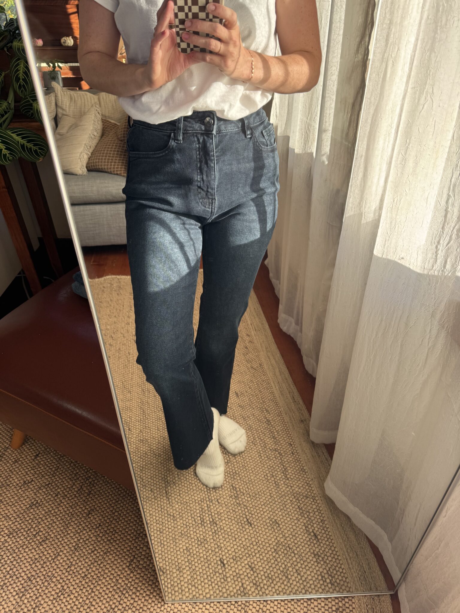Person wearing a white t-shirt, dark blue jeans, and white socks stands on a beige rug, taking a mirror selfie in a sunlit room.
