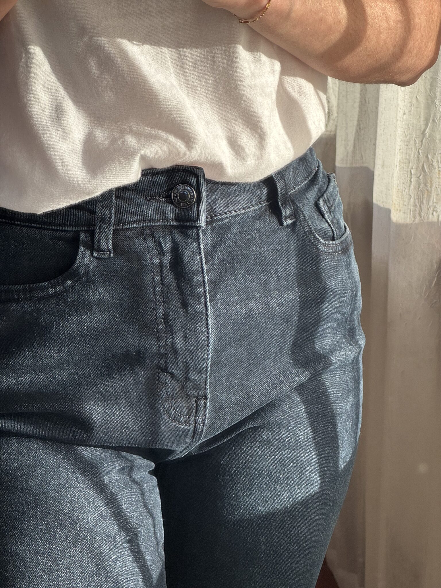 Person wearing a white t-shirt and blue jeans standing near a window with sunlight casting shadows on their clothes.