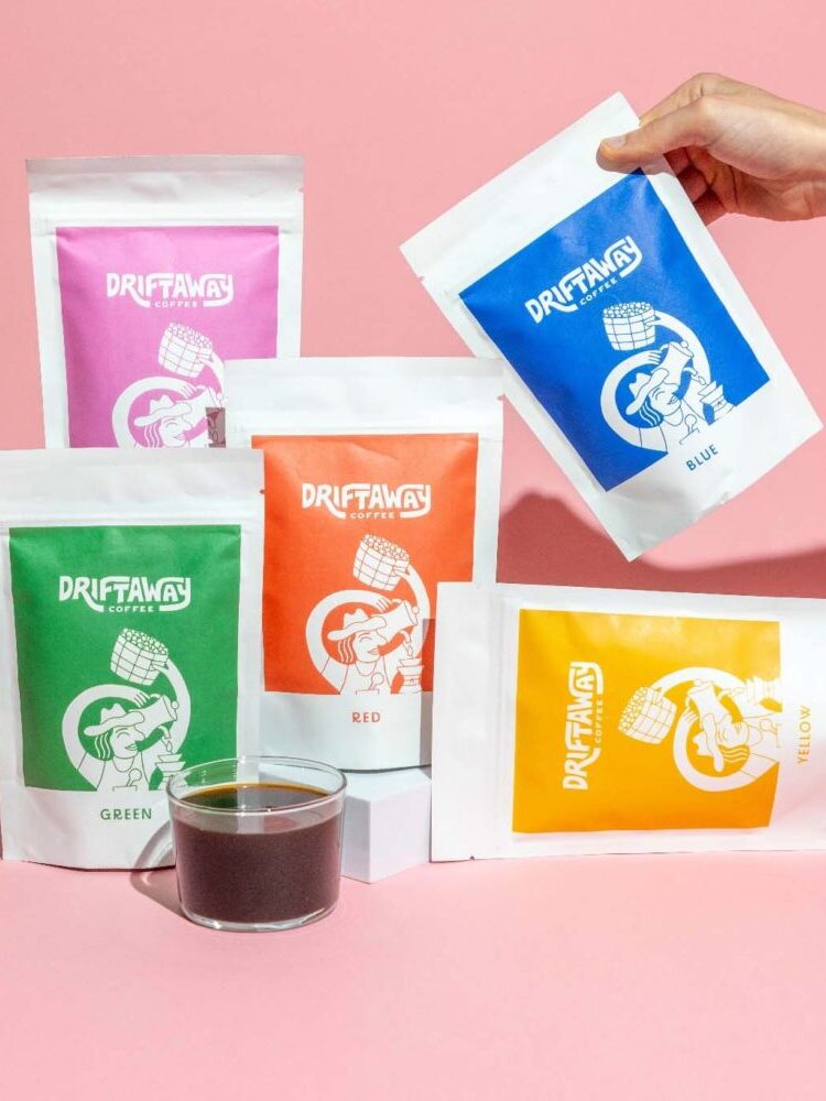 Five Driftaway Coffee bags in different colors and a glass cup of black coffee are arranged on a pink surface; a hand holds the blue bag.