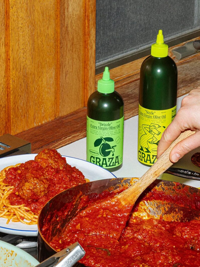 A hand stirs tomato sauce in a pan next to a plate of spaghetti and meatballs; two bottles of Graza olive oil are on the windowsill.