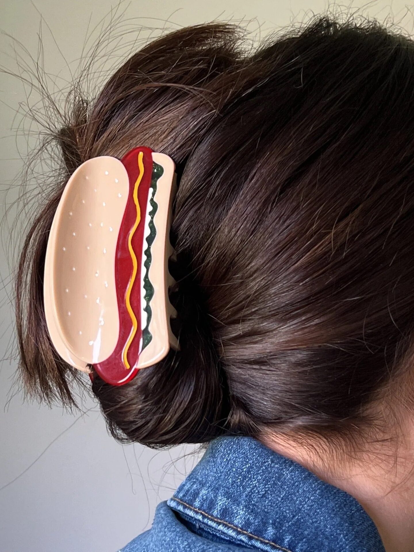 A person with brown hair wears a hair clip shaped like a hot dog with mustard, ketchup, and relish details.