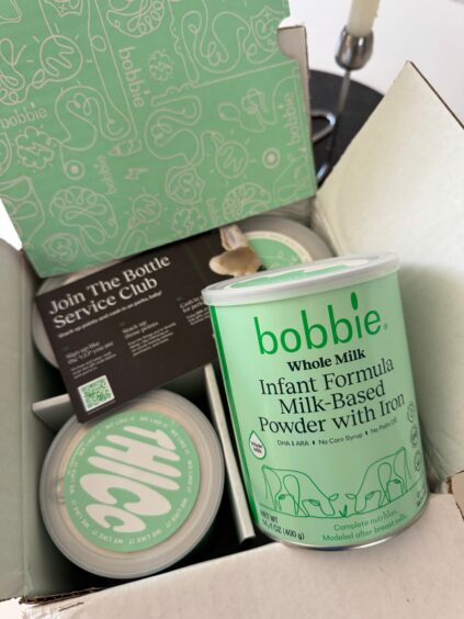 An open box containing cans of Bobbie Whole Milk Infant Formula Powder and informational card, with the packaging featuring light green branding.