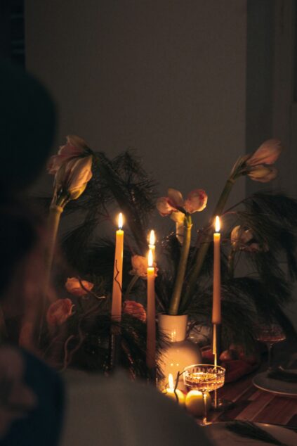 Four lit taper candles in white holders sit among flowers and greenery on a wooden table, with a blurred figure in the foreground.