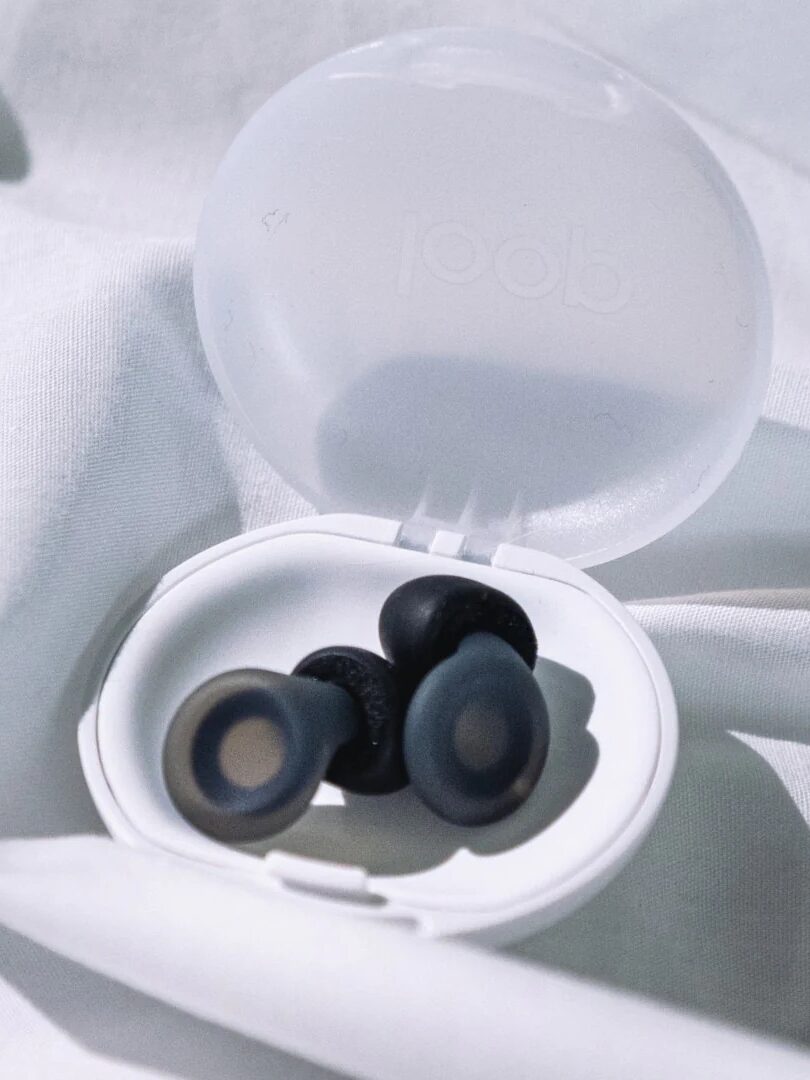 A pair of black earplugs with circular centers inside an open white plastic case, placed on a white fabric background.