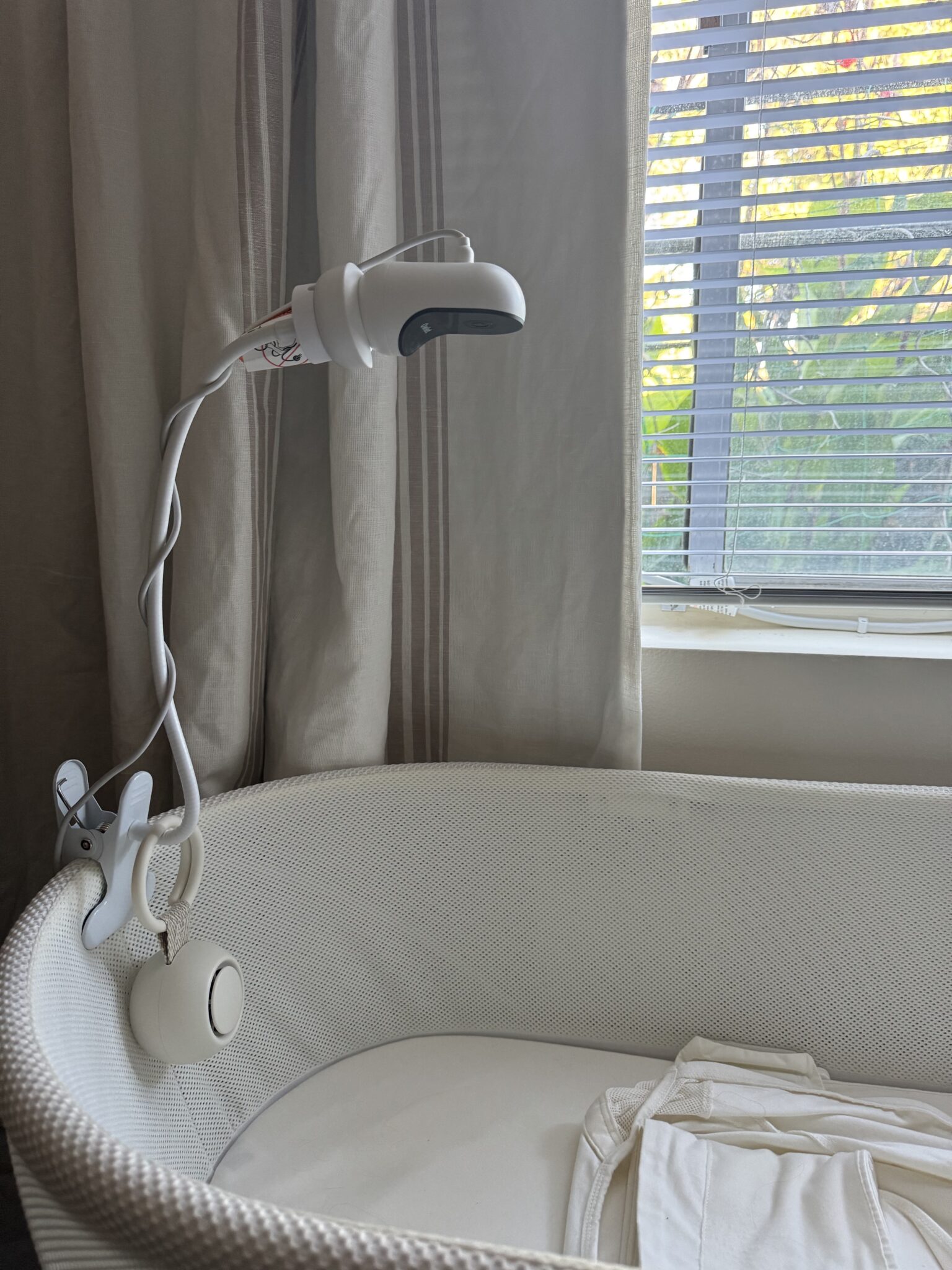 A white baby monitor camera is clipped to the side of a white bassinet near a window with closed blinds and light-colored curtains.