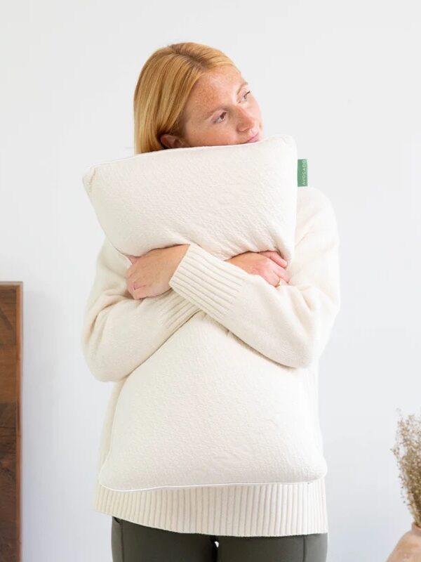A woman in a cream sweater hugs a white pillow while standing in a minimal bedroom with a wooden headboard and another pillow on the bed.