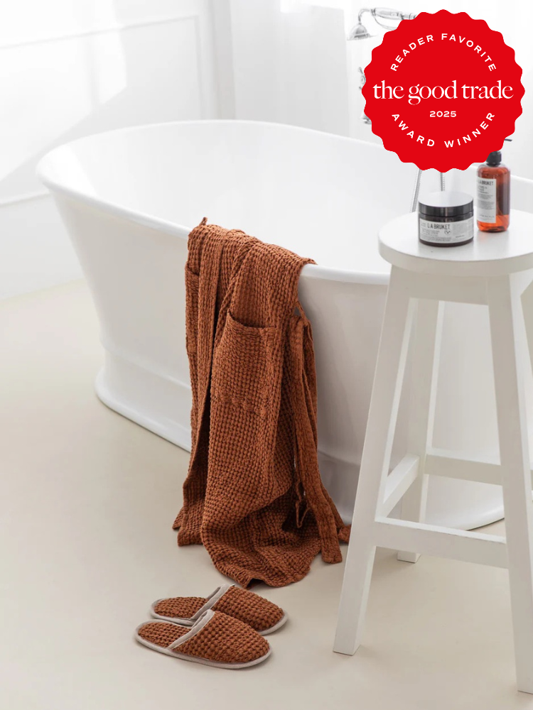 A white freestanding bathtub with a textured brown robe draped over the side, matching slippers on the floor, and bath products on a white stool. Slippers complete this serene scene, while a red award badge sits in the corner.