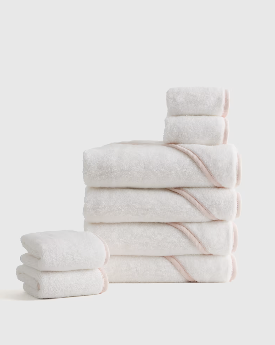 A neatly stacked set of white towels with light pink trim, including bath towels, hand towels, and washcloths, arranged against a plain background.