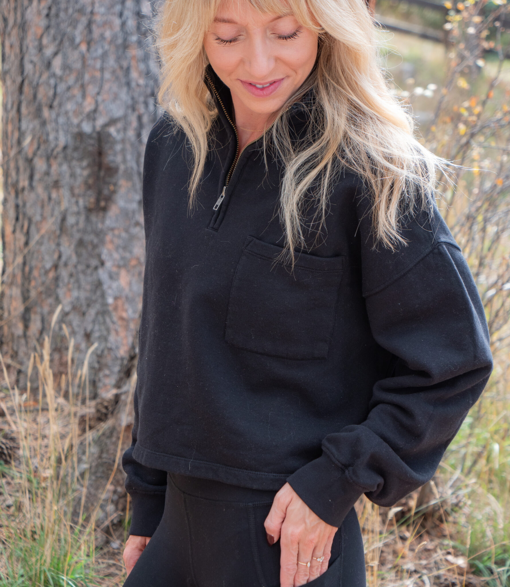 A woman with long blonde hair wearing a black quarter-zip sweatshirt and black pants stands outdoors near a tree, looking down and smiling.