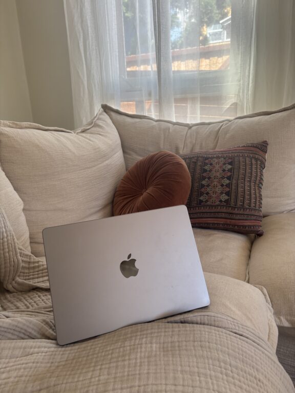 A closed silver MacBook sits on a beige couch with two decorative pillows, near a window with sheer curtains letting in natural light.