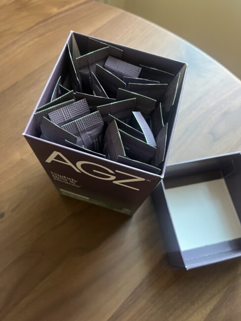 An open box labeled "AGZ" sits on a wooden table, revealing multiple individual packets inside. The box lid is placed next to it.