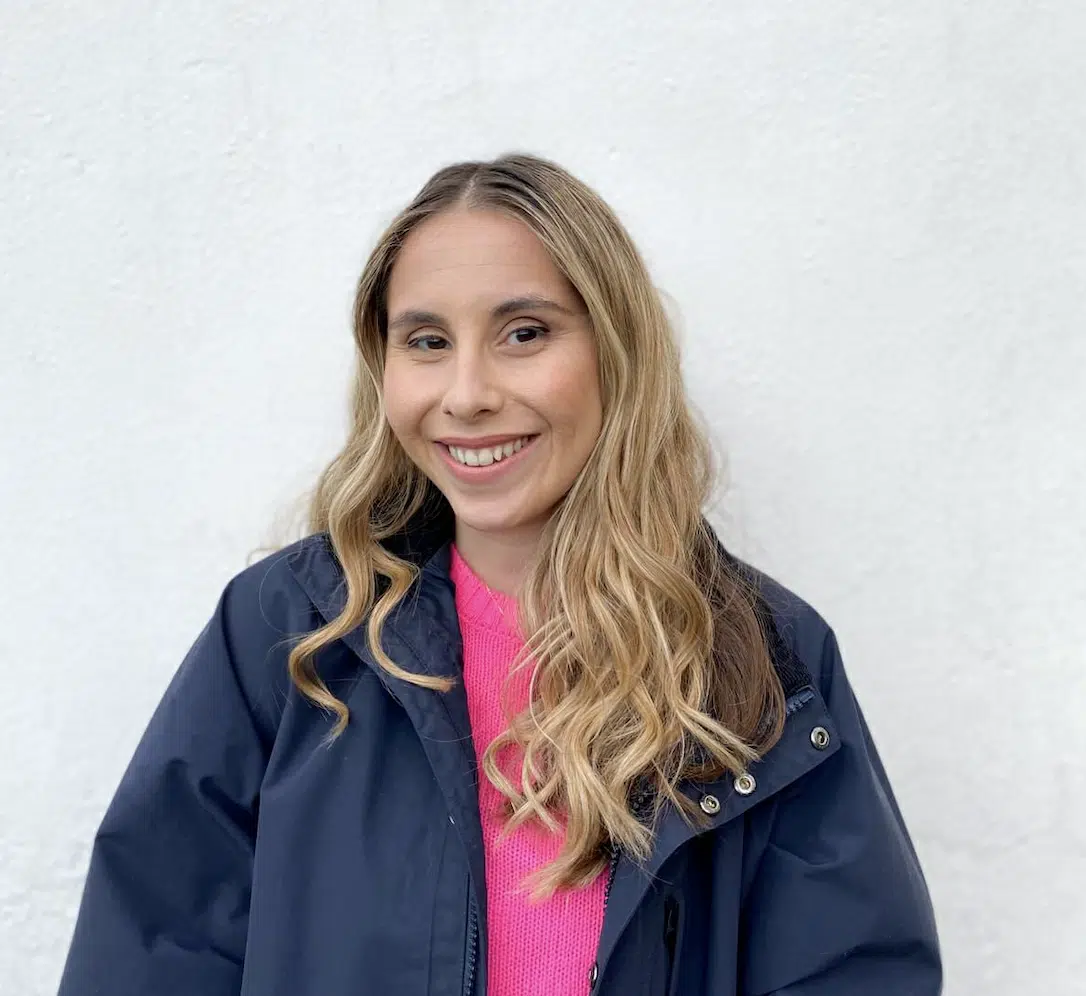 A person with long blonde hair, wearing a dark jacket over a bright pink top, stands in front of a plain white wall and smiles at the camera.