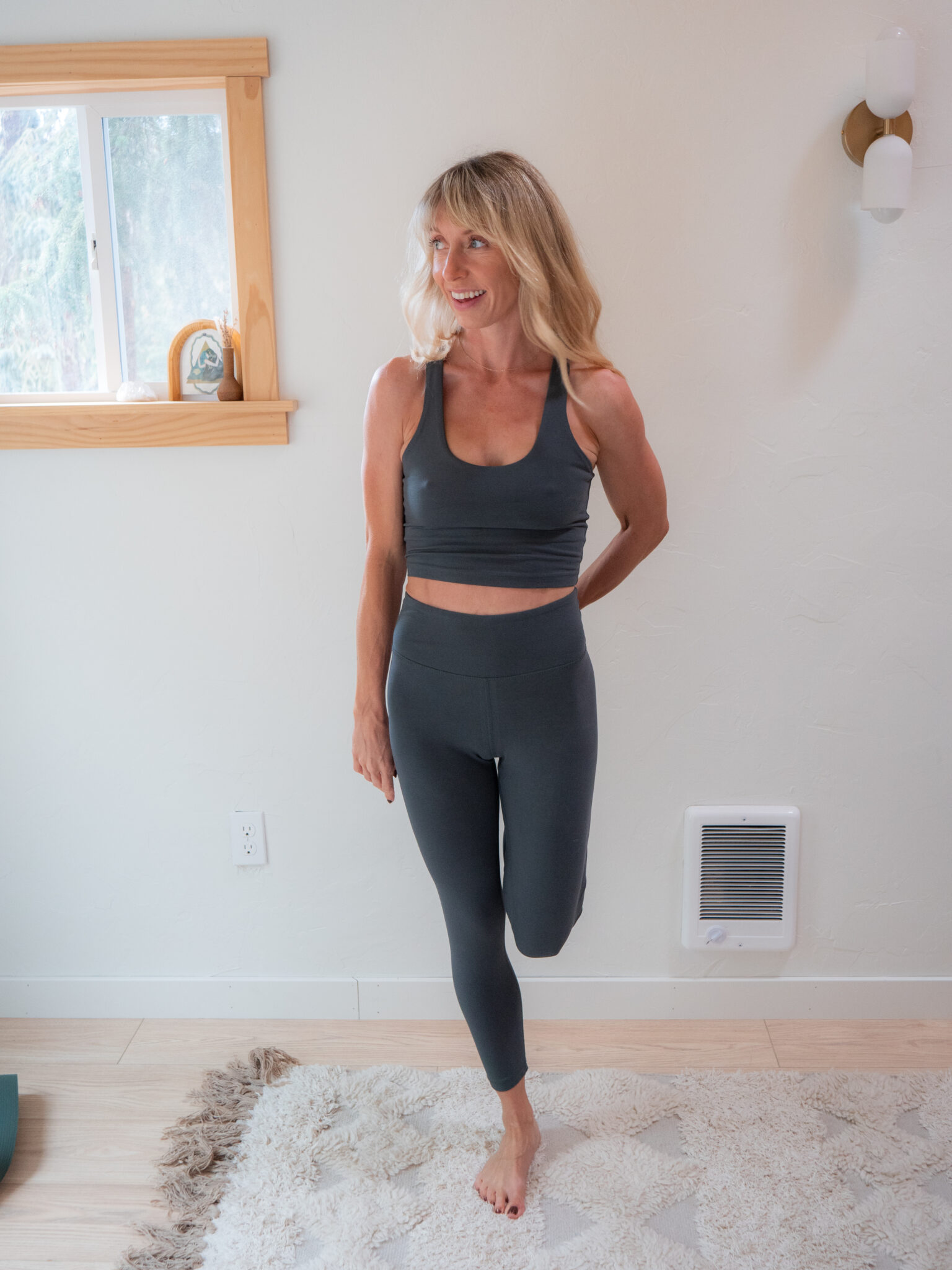 A woman in gray athletic wear stands indoors on one leg, holding her other ankle behind her in a stretching pose.