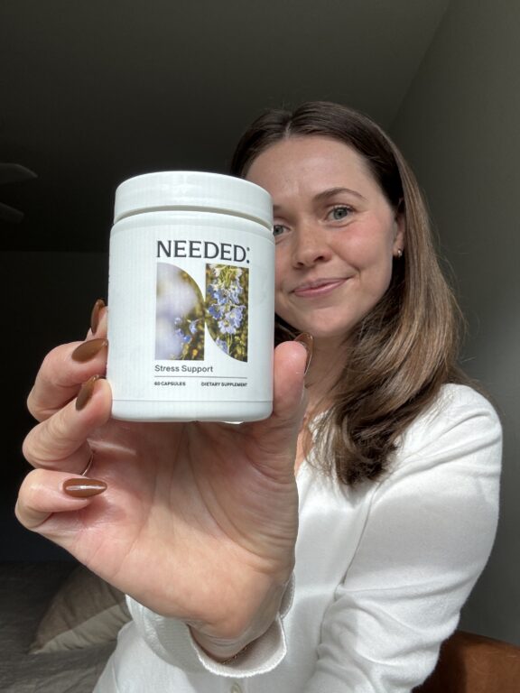 A woman in a white shirt holds up a bottle labeled “Needed: Stress Support” dietary supplement, smiling at the camera.